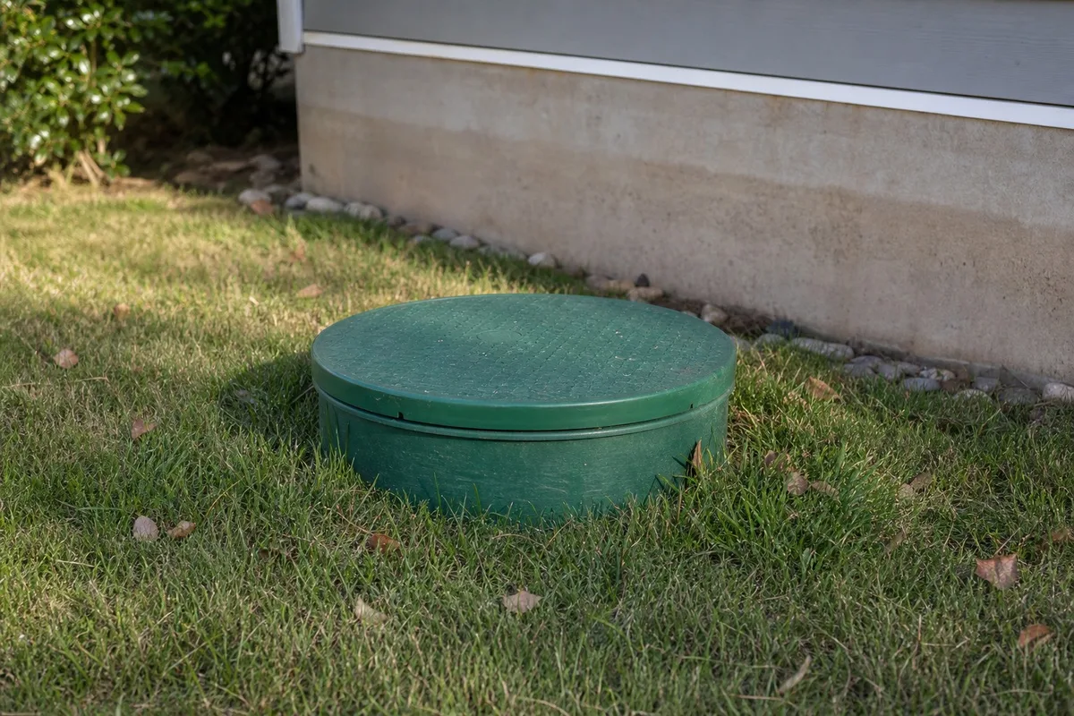 A green septic tank riser lid sits in the grass beside a home's foundation, marking the access point to an underground septic tank.