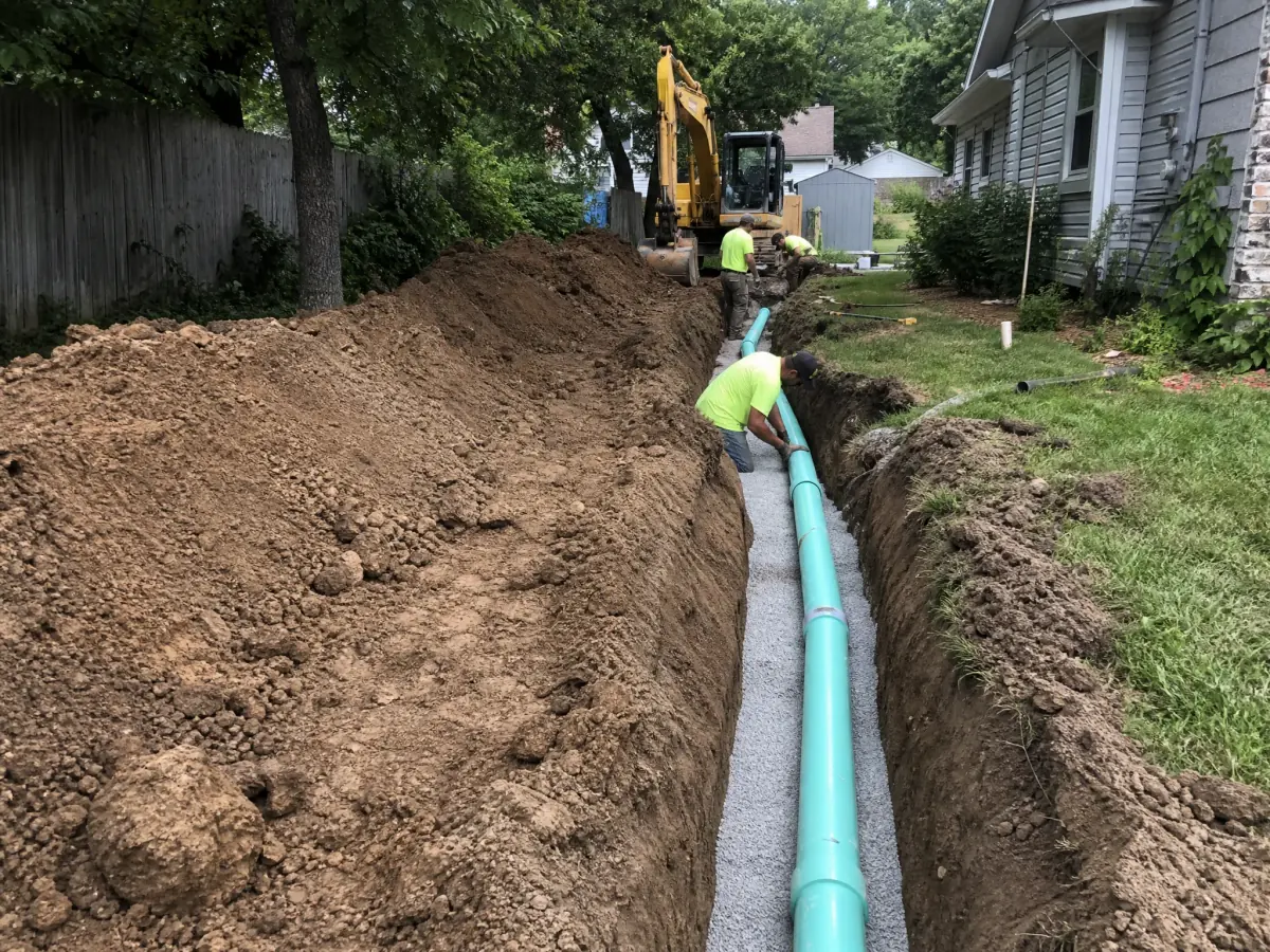 A long open trench running beside a house with a new green sewer main laid on gravel bedding, an excavator at the far end and mounded soil along the yard, showing the scale of a full line replacement.